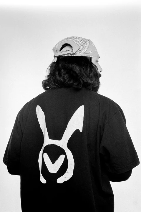 A black-and-white rear portrait showing the VAURA symbol across a tee beneath a patterned bandana