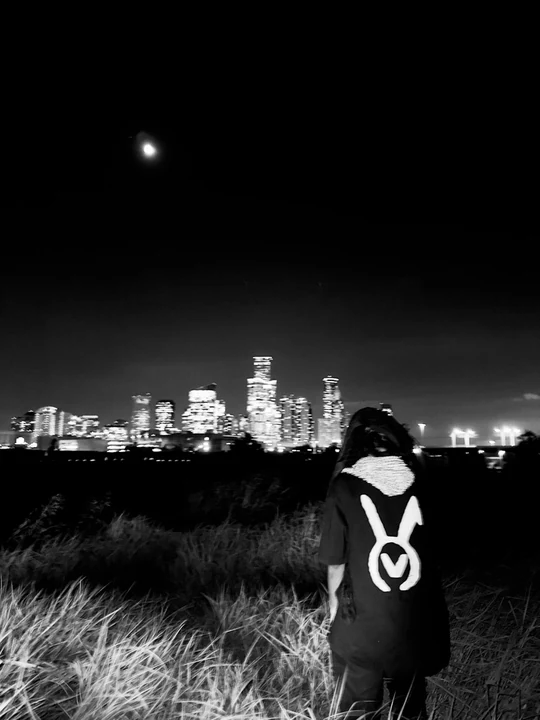 A black-and-white photograph of a person wearing the VAURA tee, looking toward an illuminated city skyline