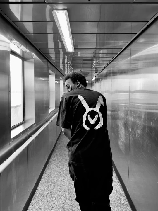 A black-and-white photograph of a person wearing the VAURA tee in a reflective corridor, its large white back symbol visible