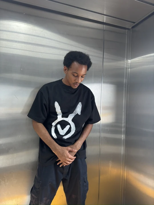 A person in a metal elevator wearing a black tee with the large white VAURA symbol facing the camera