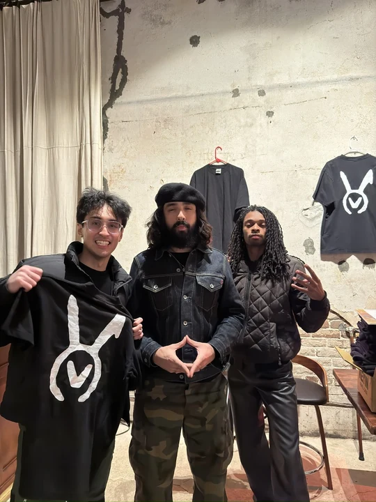 Three people at a merchandise table, one holding a black VAURA tee with the large white symbol, with another tee displayed on the wall
