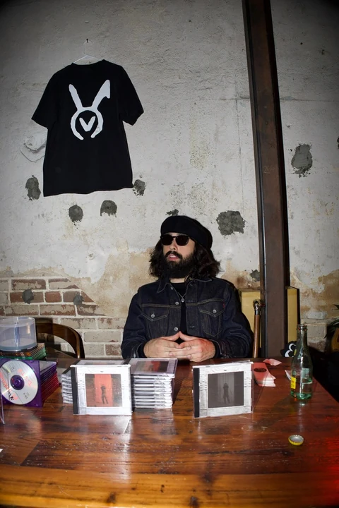A person seated behind a table of CDs with a black VAURA tee hanging on the wall
